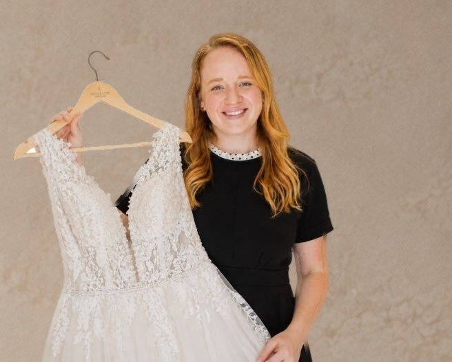 Katie, smiling, holding a lace wedding gown on a wooden hanger
